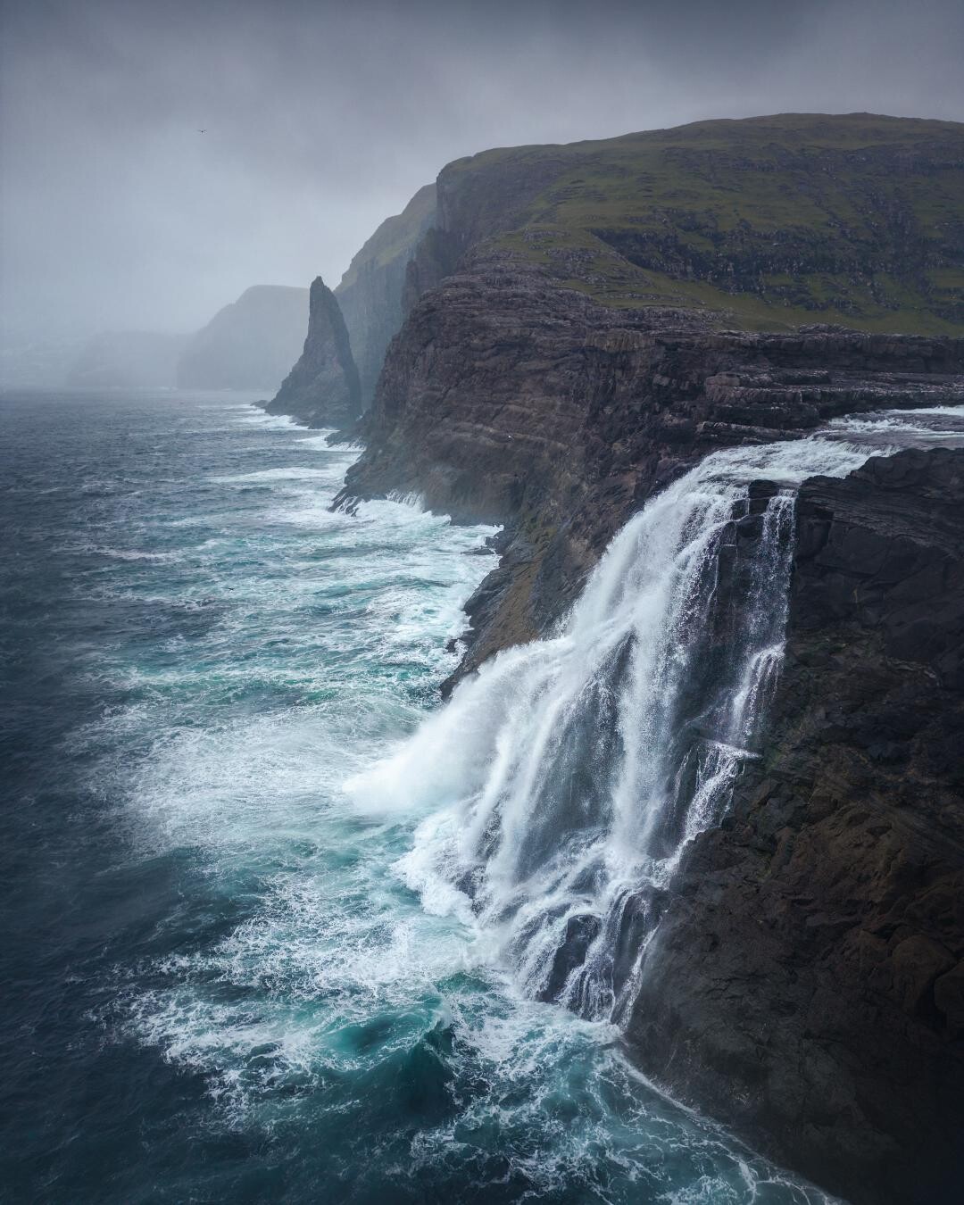 A white cascade of frothing water sweeps haphazardly down the merciless cliffs into a wild sea beneath a grim and cloudy sky. Photo credit: https://www.reddit.com/user/justerikfotos/